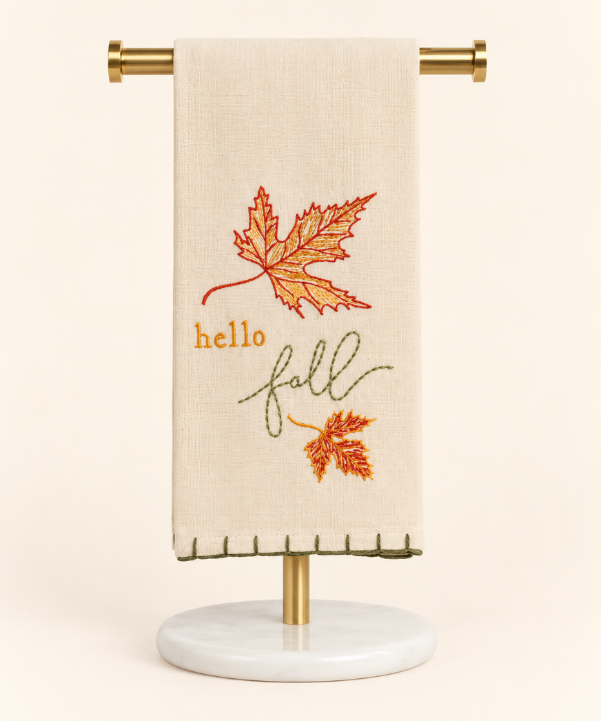 Cream embroidered “Hello Fall” hand towel with orange and gold autumn leaves displayed on a gold towel stand with a white marble base against a cream background.