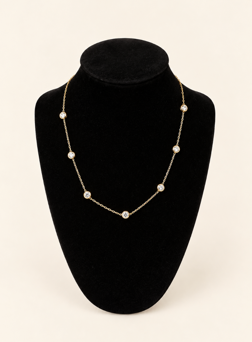 Delicate gold chain necklace featuring seven evenly spaced round bezel-set clear stones, displayed on a black velvet jewelry bust against a cream background.