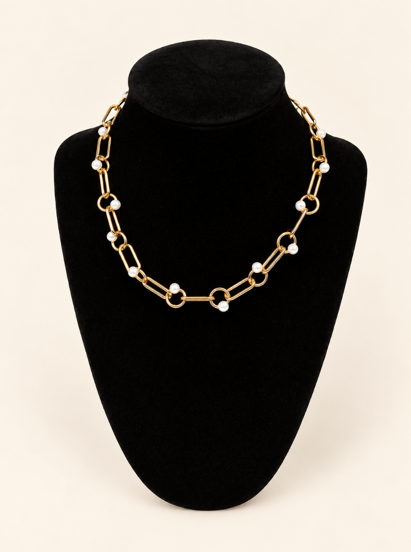 Gold statement chain necklace featuring elongated oval links, circular accents, and small white pearl details, displayed on a black velvet jewelry bust against a cream background.