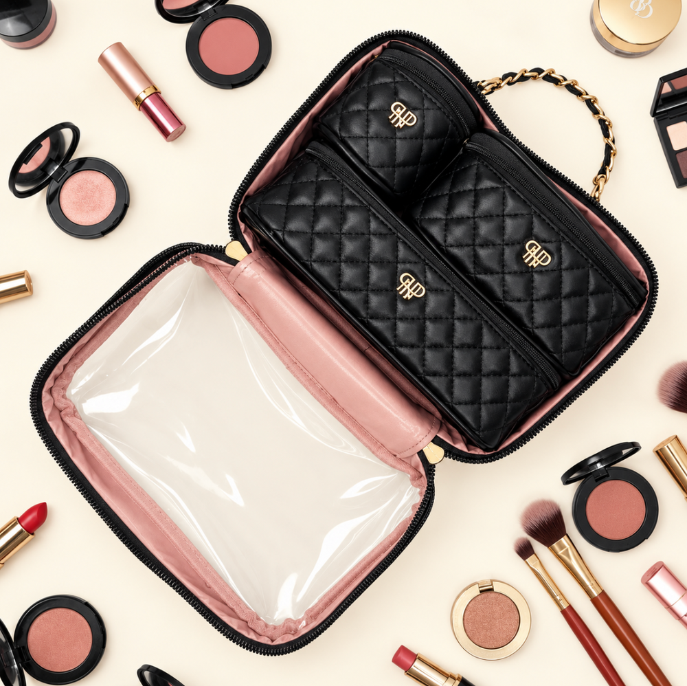 Overhead view of an open black quilted makeup travel organizer with blush pink lining, three matching zippered cosmetic pouches, and clear protective compartment, surrounded by makeup essentials on a cream background.