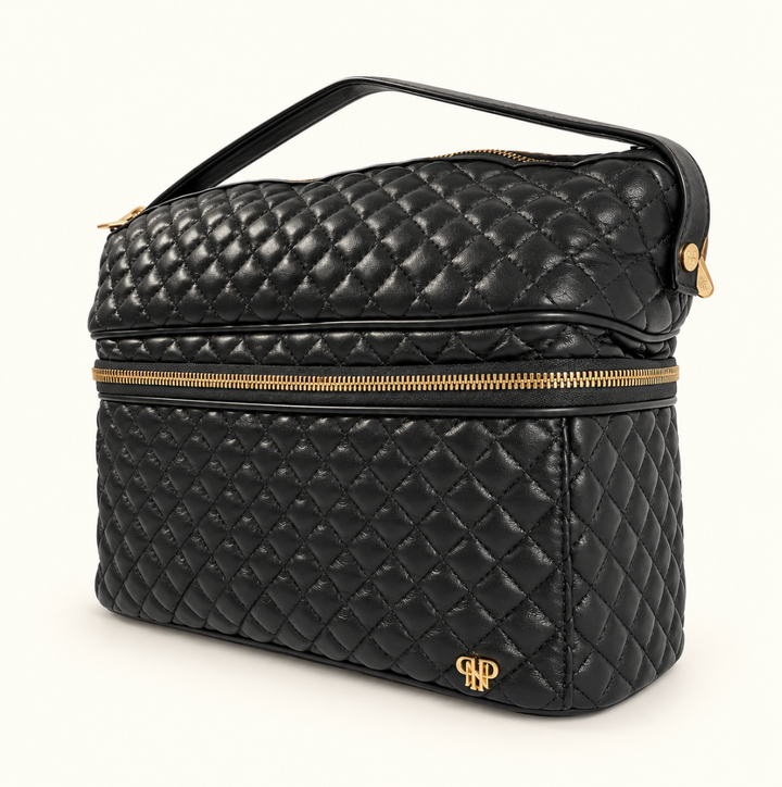 Black diamond-quilted makeup and travel organizer bag with double zippered compartments, gold hardware, structured design, and top carrying handle on a cream background.