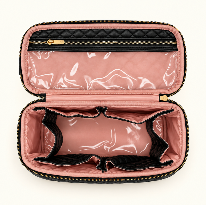 Overhead view of an open black quilted makeup travel organizer featuring blush pink wipeable lining, clear zippered storage pocket, multiple interior dividers, and gold zipper hardware on a cream background.