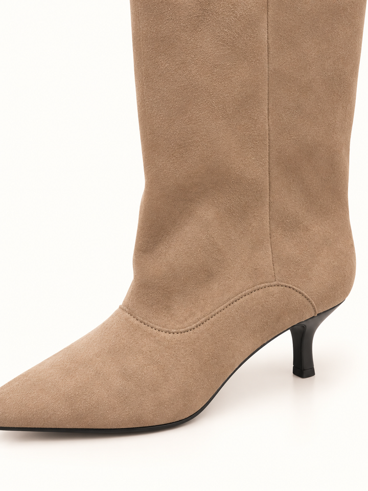 Close-up of taupe suede knee-high boot showing soft suede texture, curved ankle stitching, pointed toe, and slim black kitten heel against a cream background.