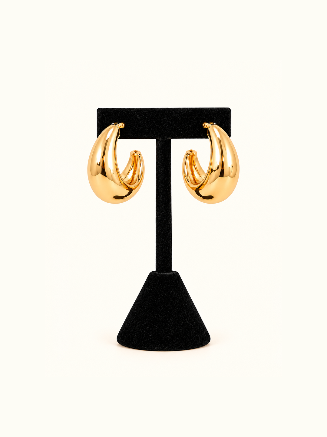 Chunky gold-tone crescent hoop earrings with a bold, sculptural curved silhouette displayed on a black velvet T-bar jewelry stand against a cream background.