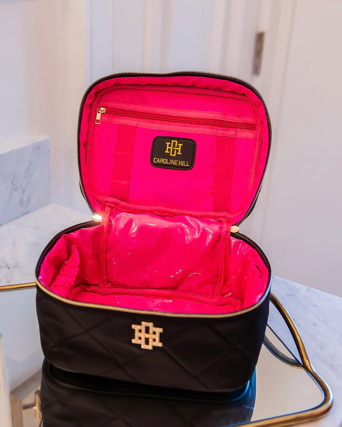 Black quilted makeup bag with pink interior, featuring a brand logo, on a reflective surface.